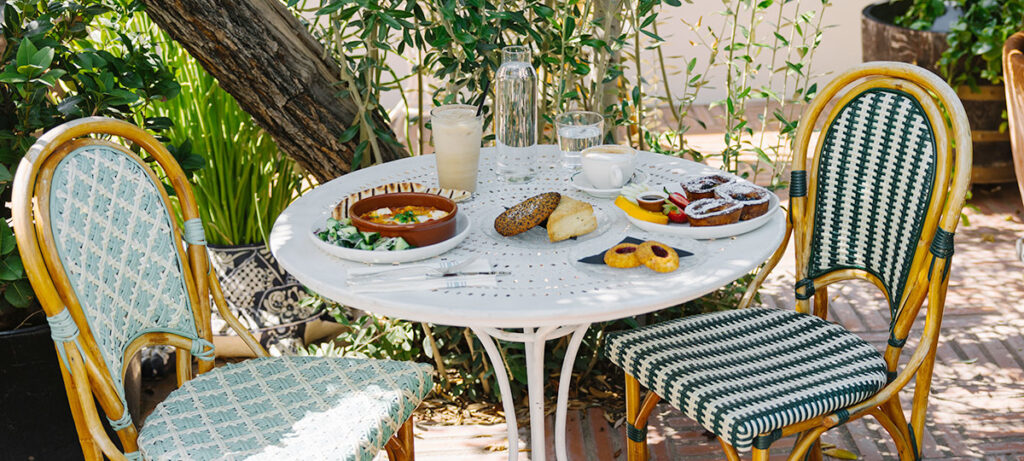 2 chairs around small table outdoors surrounded by plants. On the table there are pastries and breakfast dishes with coffees and a water pitcher