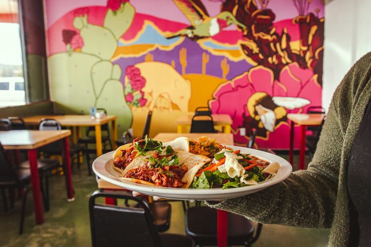 A plate of Mexican tacos being held up with a backdrop of La Chaiteria restaurant in the background